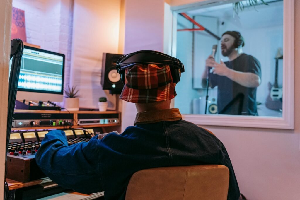 A music producer working on a recording session with an artist singing in a studio booth.